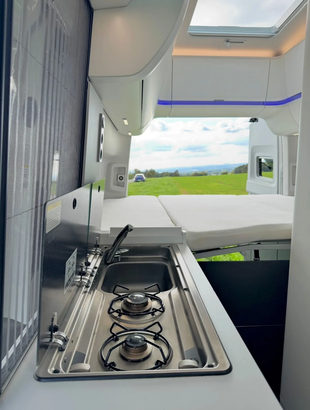 Zweiflammiger Gaskocher und Edelstahlspüle im VW Grand California mit Blick auf das Schlafbett und Panoramalandschaft